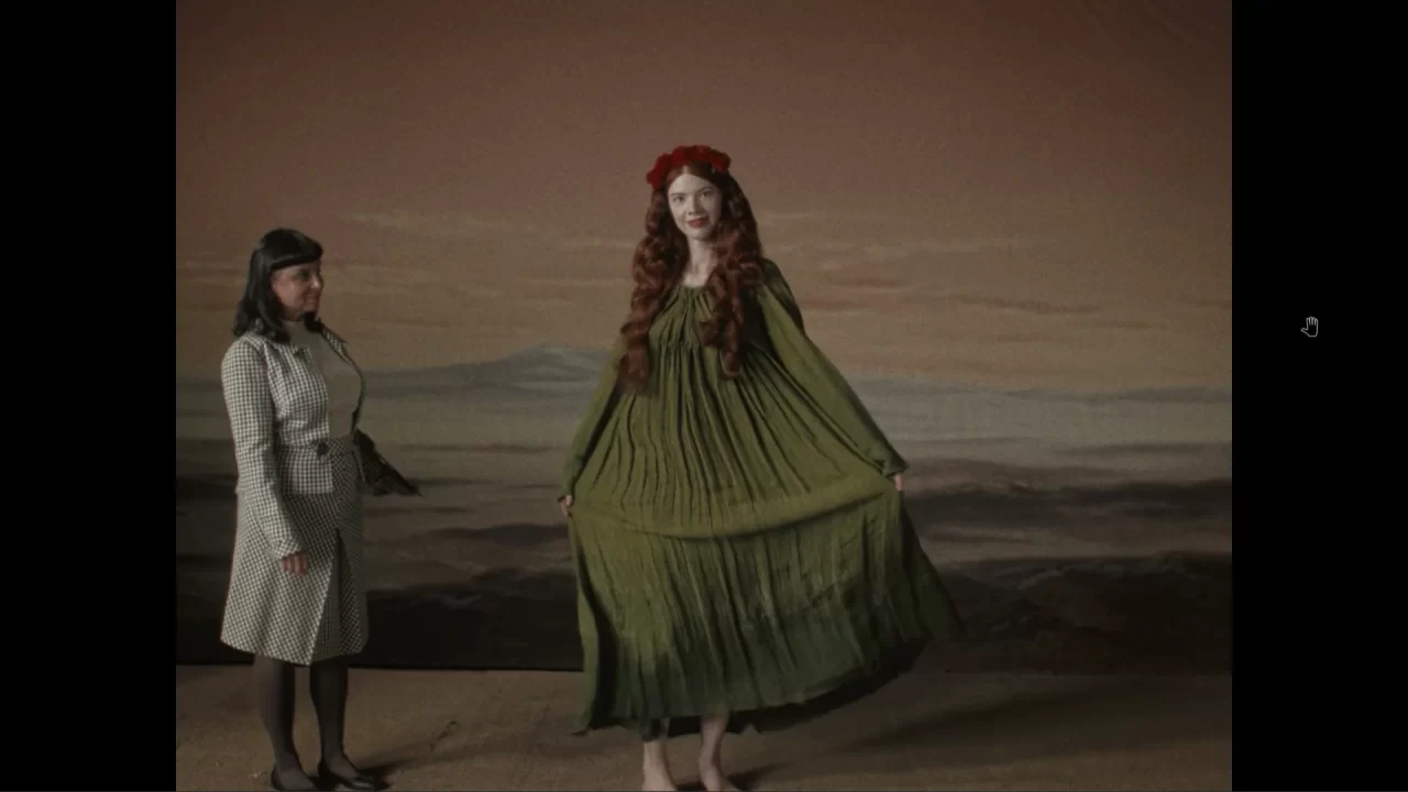 A woman with long, flowing hair and a green dress stands in front of a set background while another woman in a suit looks on.